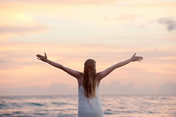 Blisstacular Living - Women with arms raised as the sun sets at the beach